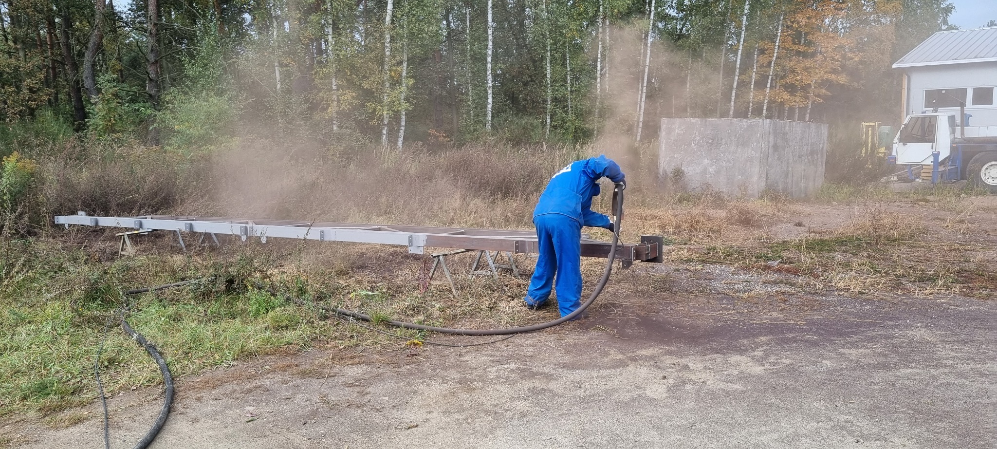 Pracownik w niebieskim kombinezonie roboczym czyści metalową konstrukcję metodą strumieniowo-ścierną na zewnątrz, unoszący się pył, w tle drzewa i budynek.