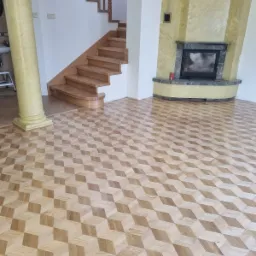 Parkiet ułożony we wzór 3D kostek w jasnym salonie z drewnianymi schodami i kominkiem z zielonego marmuru.