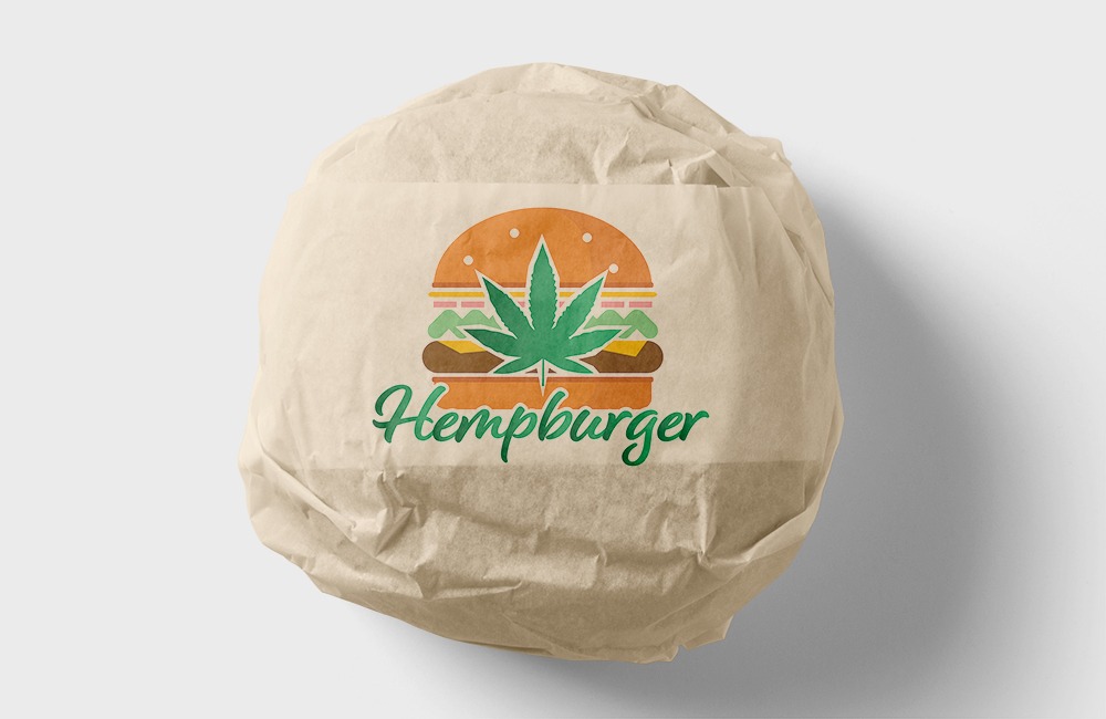 Grafika logo Hempburger na papierowej torbie, przedstawiająca burgera z liściem konopi zamiast mięsa, w zielonej kolorystyce.