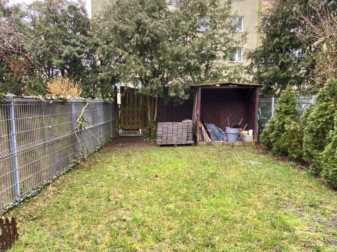 Widok ogrodu przed realizacją prac: zaniedbany trawnik, metalowe ogrodzenie z fragmentami uschniętej roślinności, drewniana szopa z narzędziami i stosem kostki brukowej w tle.
