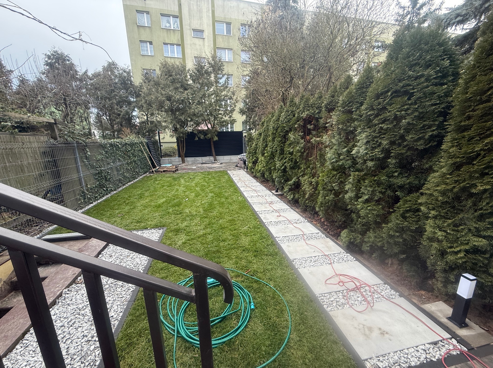 Świeżo położona trawa z rolki w ogrodzie miejskim, widok z góry na ścieżkę z płyt betonowych z białym grysem, obok zwinięty wąż ogrodowy i rząd tuj.