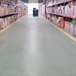 Długi korytarz magazynowy z regałami wypełnionymi kartonami i pudełkami, wyznaczony żółtymi liniami na podłodze, z wózkiem transportowym w oddali.