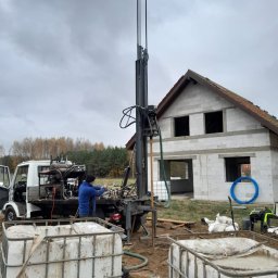 Z.U.H.P ŁUKASZ TOMASIK - Wiercenie studni przy nowo powstającym domu z pustaków; widoczna ciężarówka z wiertnicą, pracownik w niebieskiej bluzie obsługujący maszynę, białe zbiorniki na wodę i szary, pochmurny dzień.