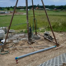 Z.U.H.P ŁUKASZ TOMASIK - Wiertnica do studni głębinowej na placu budowy, widoczna rura z niebiesko-zielonymi oznaczeniami leżąca na ziemi obok nowo ułożonej ażurowej kostki brukowej.