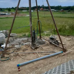Wiertnica do studni głębinowej na placu budowy, widoczna rura z niebiesko-zielonymi oznaczeniami leżąca na ziemi obok nowo ułożonej ażurowej kostki brukowej.