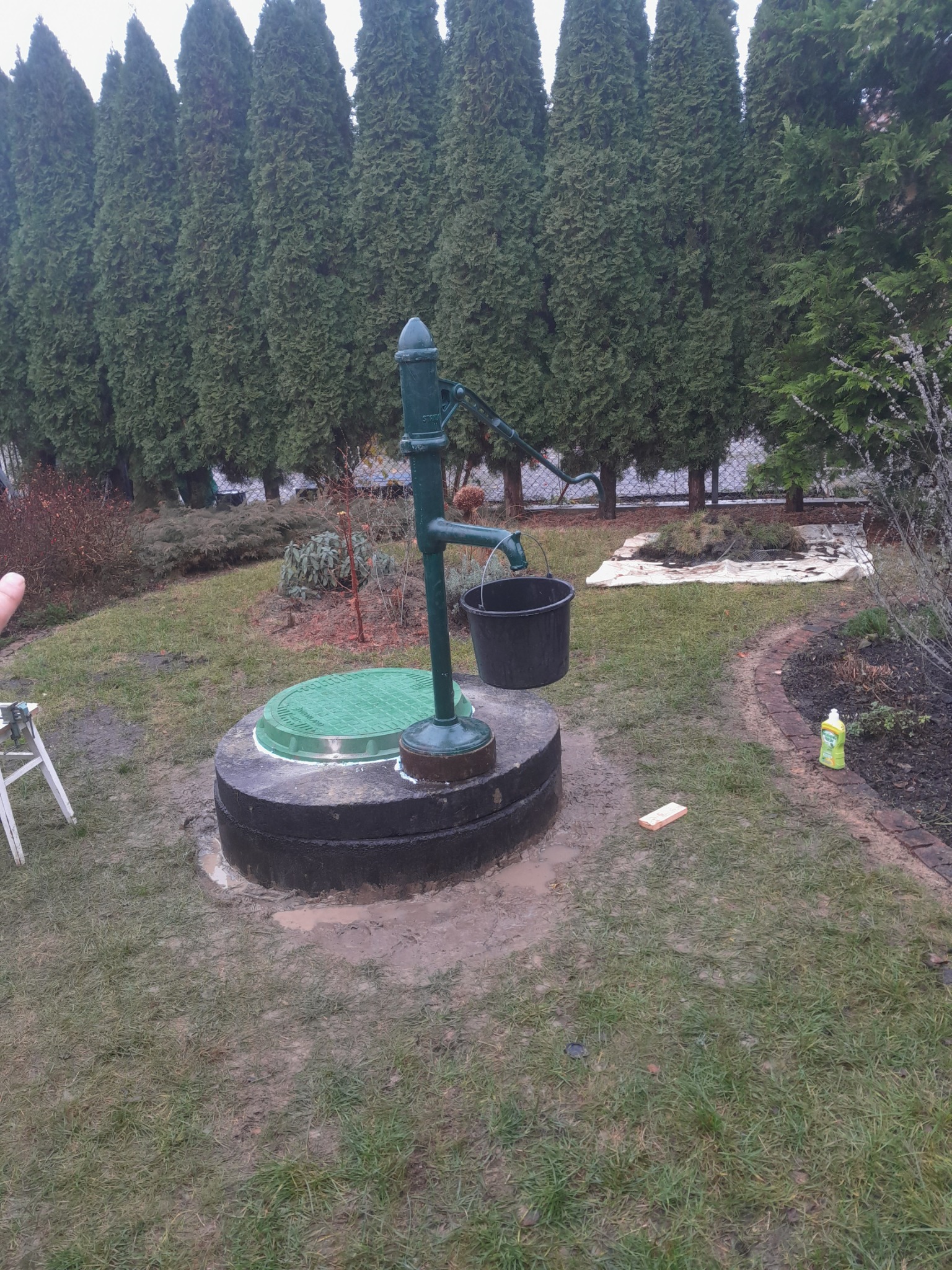 Zakończona budowa studni kręgowej z zieloną pokrywą i ręczną pompą w ogrodzie, widoczny czarny kubeł, tło stanowią wysokie tuje.