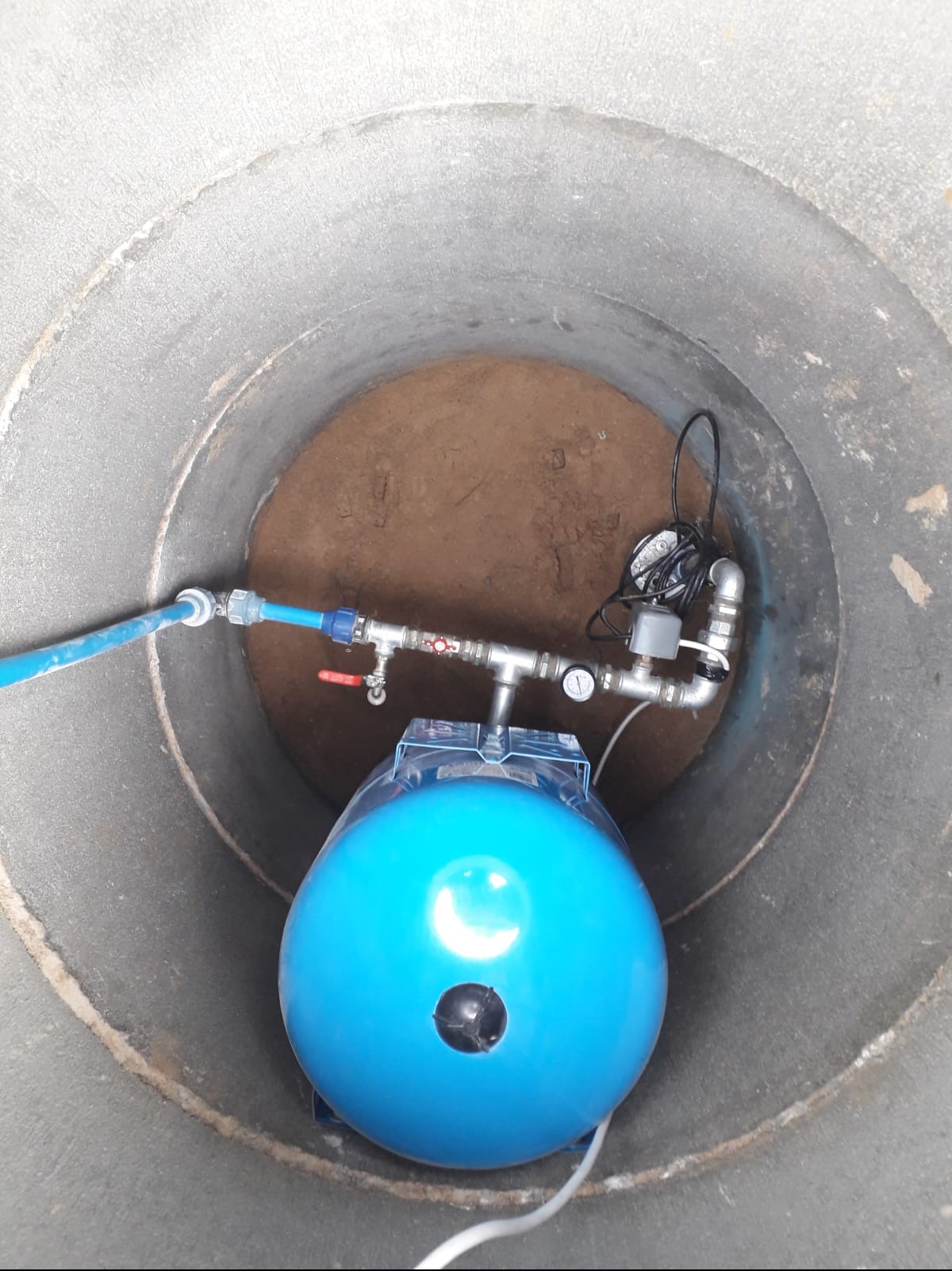Wnętrze betonowej studni kręgowej z zainstalowanym hydroforem w kolorze niebieskim, rurami, manometrem i elementami elektrycznymi. Na dnie widoczna ziemia.