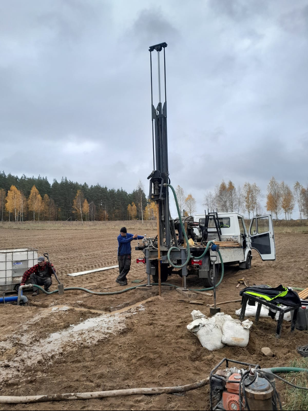 Wiertnica zamontowana na samochodzie podczas pracy w polu, dwóch pracowników obsługuje maszynę, jeden z nich w czerwonej koszuli w kratę przyłącza rury, drugi w niebieskiej bluzie kontroluje pracę...