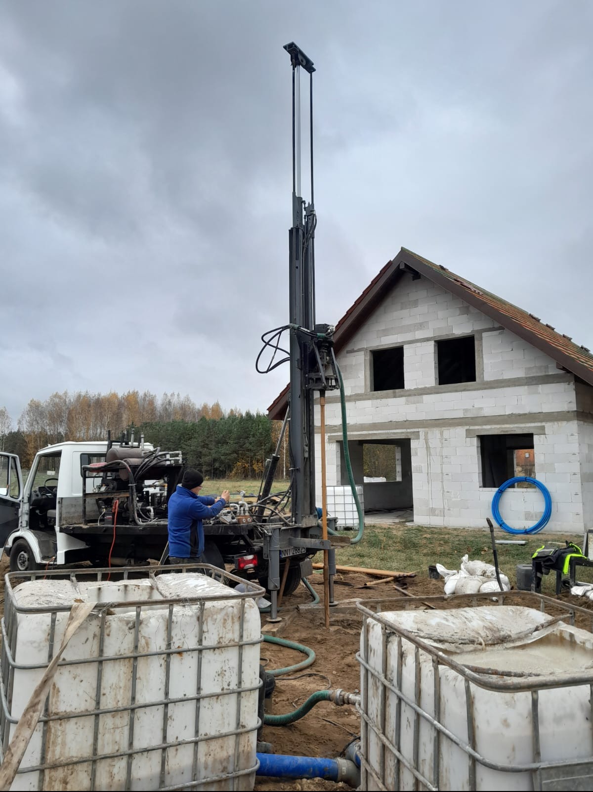 Wiercenie studni przy nowo powstającym domu z pustaków; widoczna ciężarówka z wiertnicą, pracownik w niebieskiej bluzie obsługujący maszynę, białe zbiorniki na wodę i szary, pochmurny dzień.