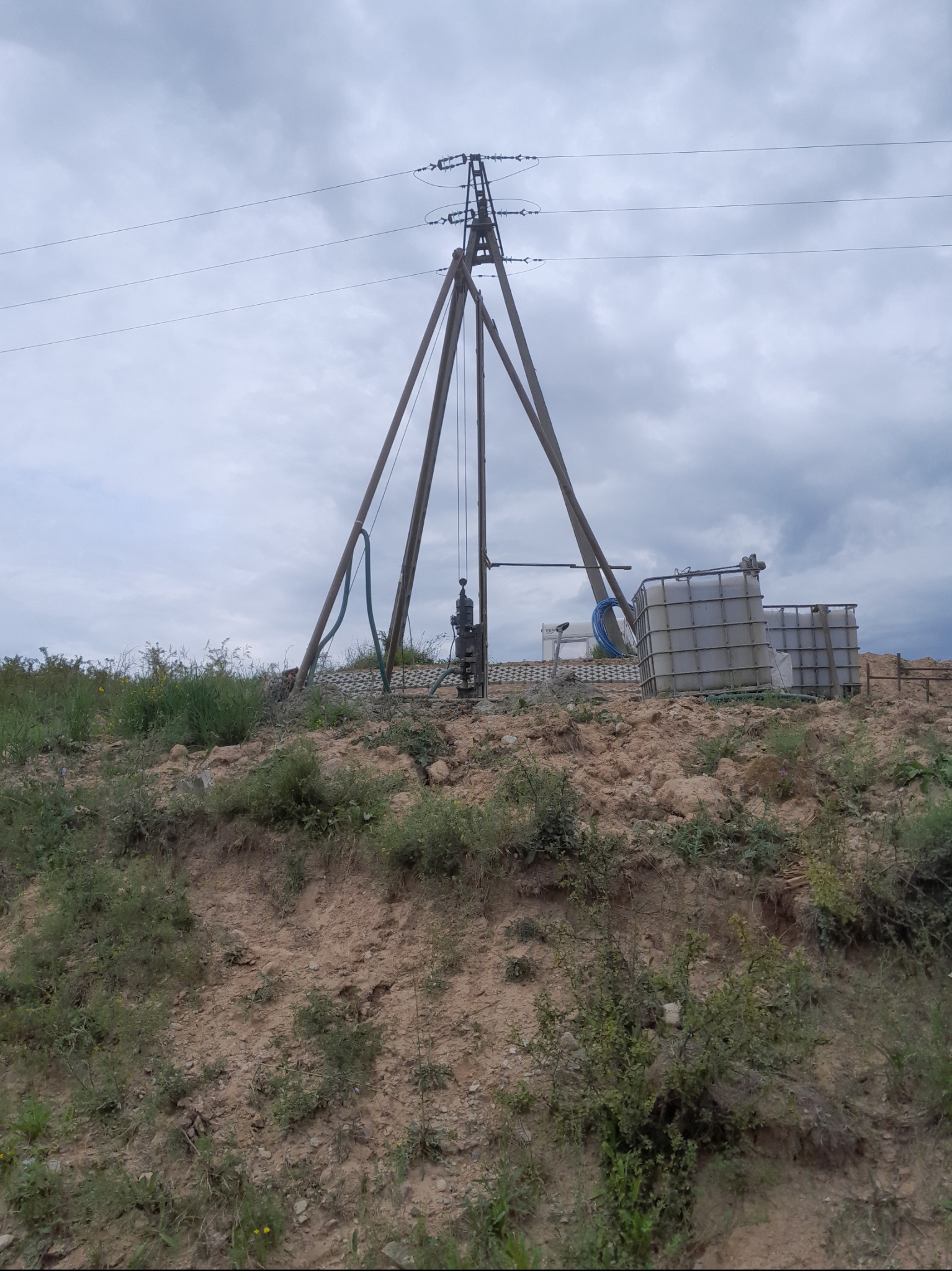 Drewniana konstrukcja wiertnicza z osprzętem na wzgórzu, obok zbiorniki na wodę, linia wysokiego napięcia w tle pod pochmurnym niebem.