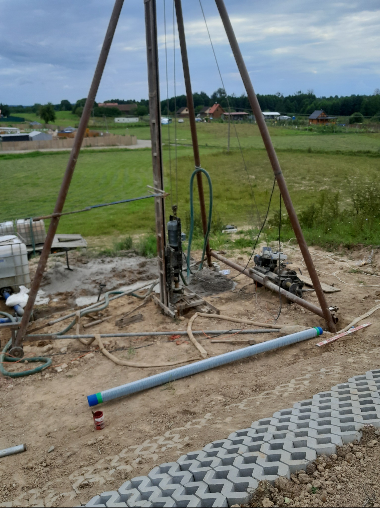 Wiertnica do studni głębinowej na placu budowy, widoczna rura z niebiesko-zielonymi oznaczeniami leżąca na ziemi obok nowo ułożonej ażurowej kostki brukowej.