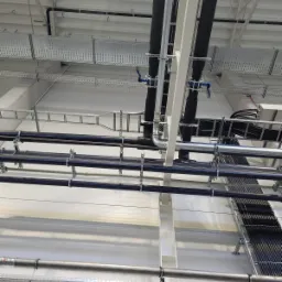Rzut z dołu na sufit hali przemysłowej z licznymi rurami, przewodami i korytami kablowymi, niektóre rury izolowane, z widocznymi zaworami kulowymi z niebieskimi uchwytami.