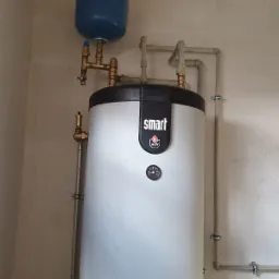 Bojler ACV Smart z niebieskim naczyniem wzbiorczym, widoczne rury i instalacja, ściana z notatkami w tle.