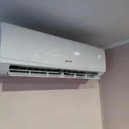 Biała klimatyzacja Sinclair zamontowana na różowej ścianie w rogu pomieszczenia, wyświetlająca temperaturę 19 stopni.