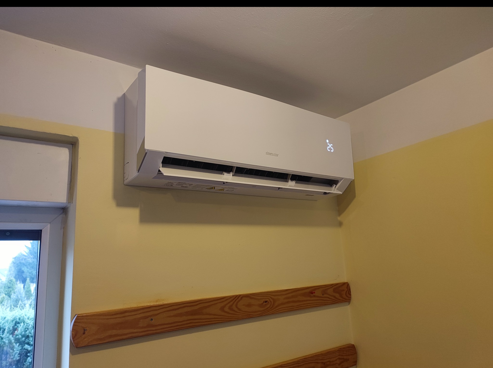 Biały klimatyzator marki Sinclair zamontowany na żółtej ścianie w pomieszczeniu, widoczne drewniane listwy przypodłogowe i fragment okna.