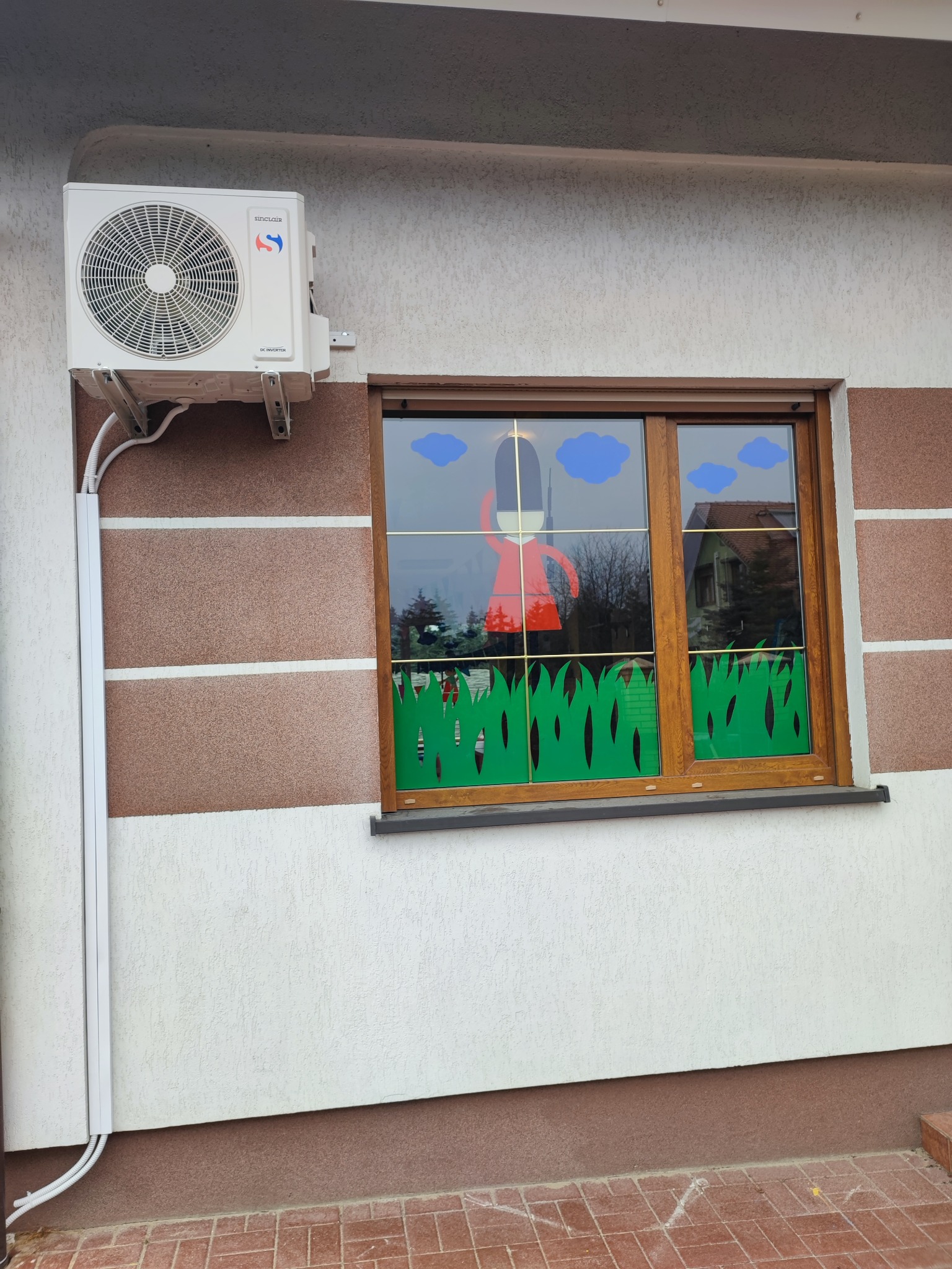 Biała jednostka zewnętrzna klimatyzacji Sinclair zamontowana na ścianie domu z dekoracją okna przedstawiającą żołnierza i chmury.