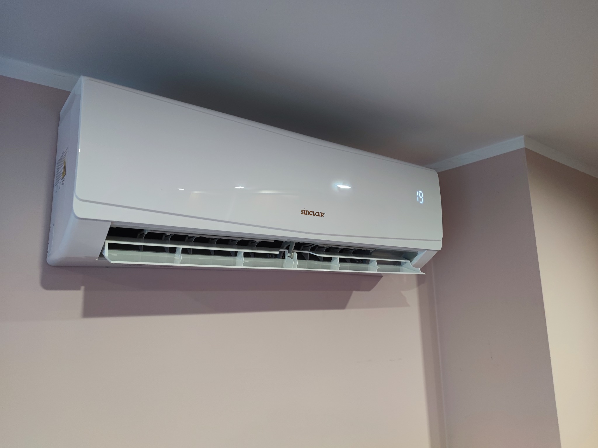 Biała klimatyzacja Sinclair zamontowana na różowej ścianie w rogu pomieszczenia, wyświetlająca temperaturę 19 stopni.