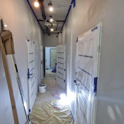 ODNOWA-Uslugi Remontowe Ihor Chornyi - Korytarz w trakcie malowania, zabezpieczone folią drzwi, widoczna drabina, taśma malarska na suficie i ścianach, podłoga przykryta folią ochronną.