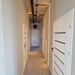 ODNOWA-Uslugi Remontowe Ihor Chornyi - Nowoczesny korytarz z szarą podłogą z płytek, białymi drzwiami z czarnymi paskami, oświetleniem LED w pionowych wnękach i surowym betonowym sufitem z widoczną instalacją elektryczną.