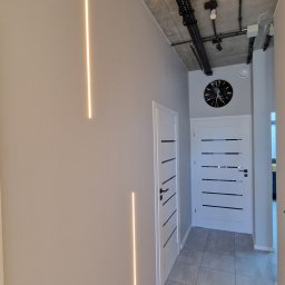 ODNOWA-Uslugi Remontowe Ihor Chornyi - Nowoczesny korytarz z podświetlanymi pionowymi listwami LED na szarej ścianie, betonowy sufit z widocznymi instalacjami, dwie białe drzwi z czarnymi akcentami i duży zegar na ścianie.