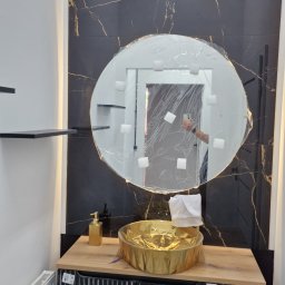 ODNOWA-Uslugi Remontowe Ihor Chornyi - Nowoczesna łazienka z okrągłym lustrem w folii ochronnej, złotą umywalką i czarnymi płytkami imitującymi marmur ze złotymi żyłami.