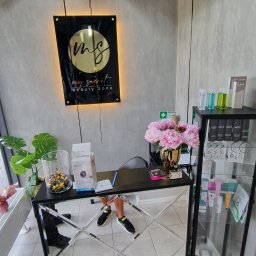 ODNOWA-Uslugi Remontowe Ihor Chornyi - Recepcja w salonie kosmetycznym z czarnym biurkiem z lustrzanymi wstawkami, na którym stoi wazon z różowymi piwoniami, obok witryna z kosmetykami, na ścianie podświetlane logo salonu.