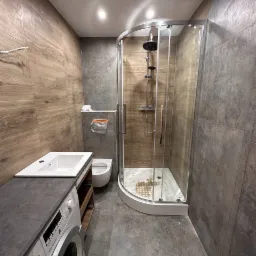 Nowoczesna łazienka z kabiną prysznicową typu walk-in, umywalką nablatową z szarą szafką i pralką Bosch Serie 4, wykończona płytkami imitującymi drewno i szarym betonem.