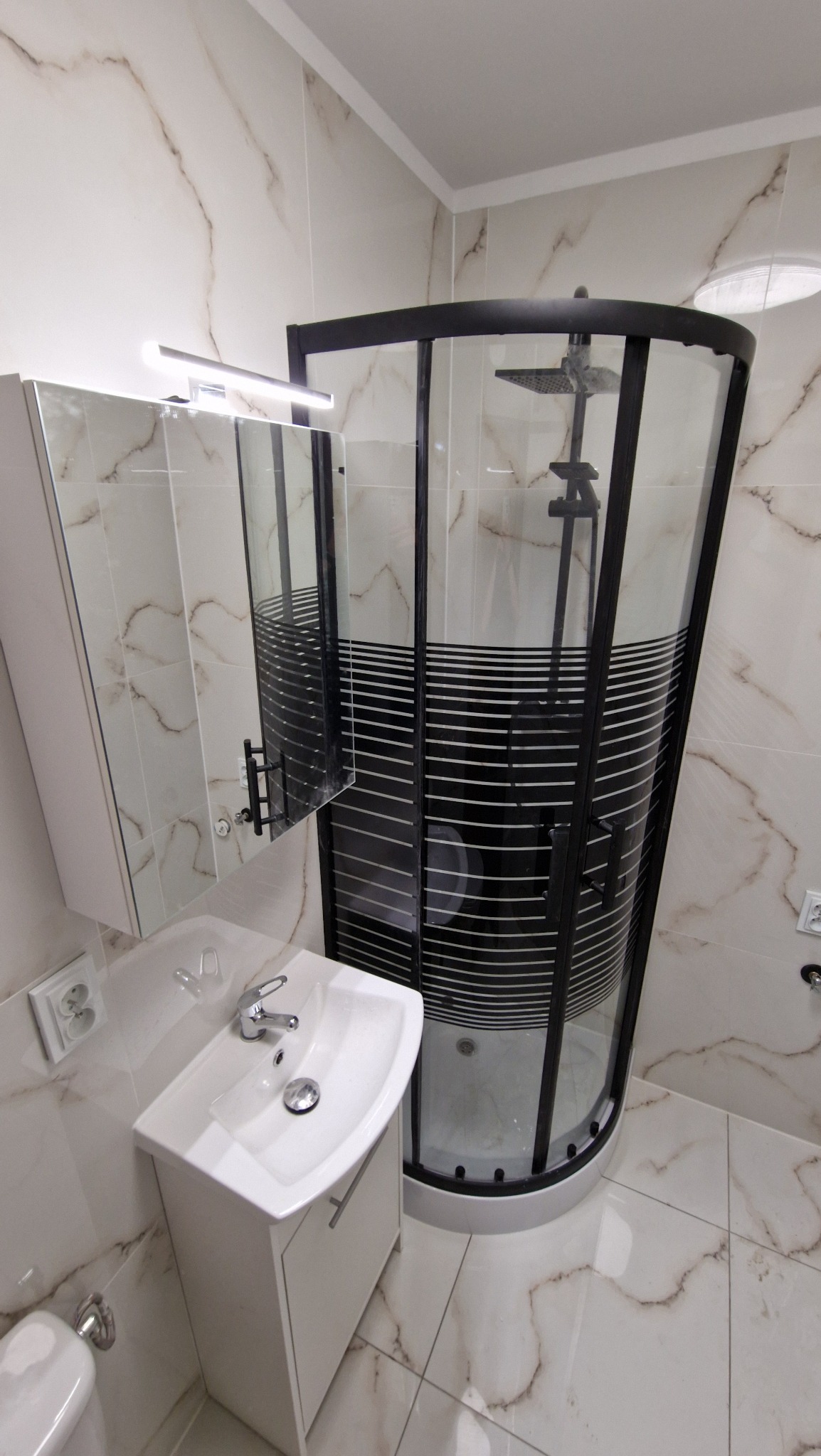 Nowoczesna łazienka z kabiną prysznicową z czarnymi profilami i poziomymi pasami, umywalką z szafką w kolorze białym oraz lustrem z oświetleniem, wykończona płytkami imitującymi marmur.