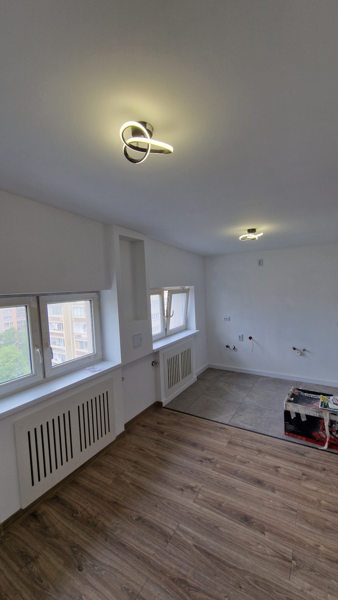 Wnętrze pokoju w trakcie prac wykończeniowych: położona podłoga z paneli, częściowo ułożone płytki, białe ściany z wyprowadzonymi instalacjami, grzejniki pod oknami, nowoczesne lampy sufitowe.