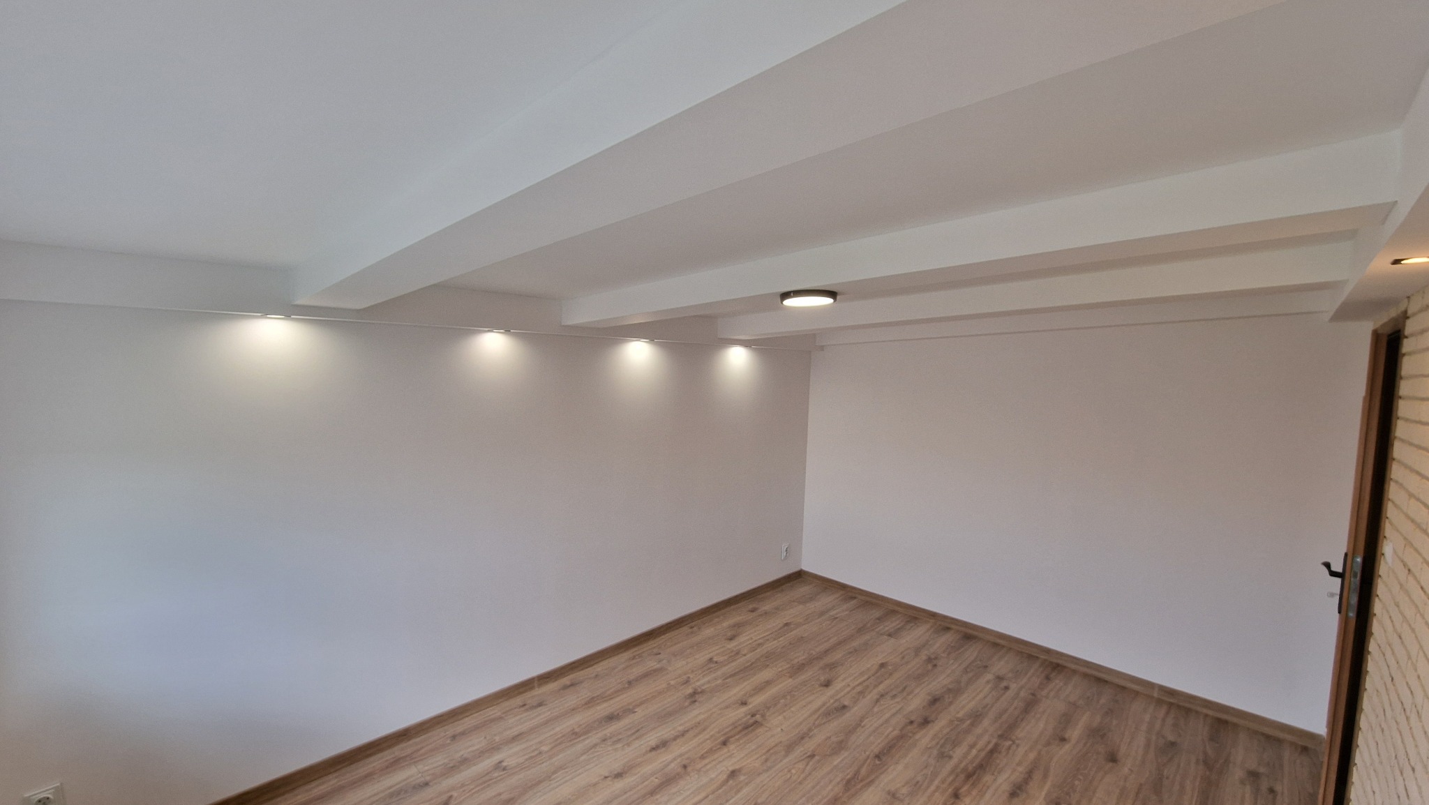 Pusty pokój po remoncie z panelami podłogowymi, białymi ścianami, listwami przypodłogowymi, sufitem z belkami i oświetleniem punktowym.