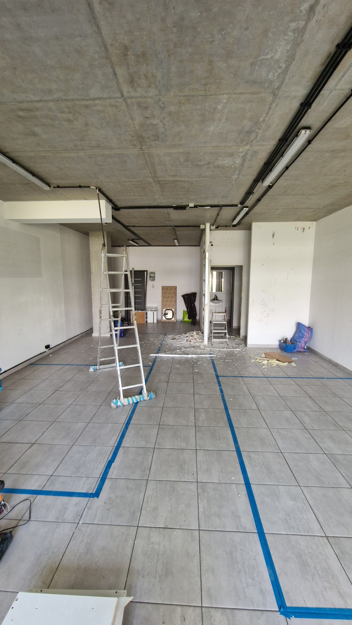 Przestrzeń w trakcie demontażu ścian działowych, widoczne gruz i zabezpieczona podłoga z płytek, drabina przystawna z zabezpieczeniem antypoślizgowym.