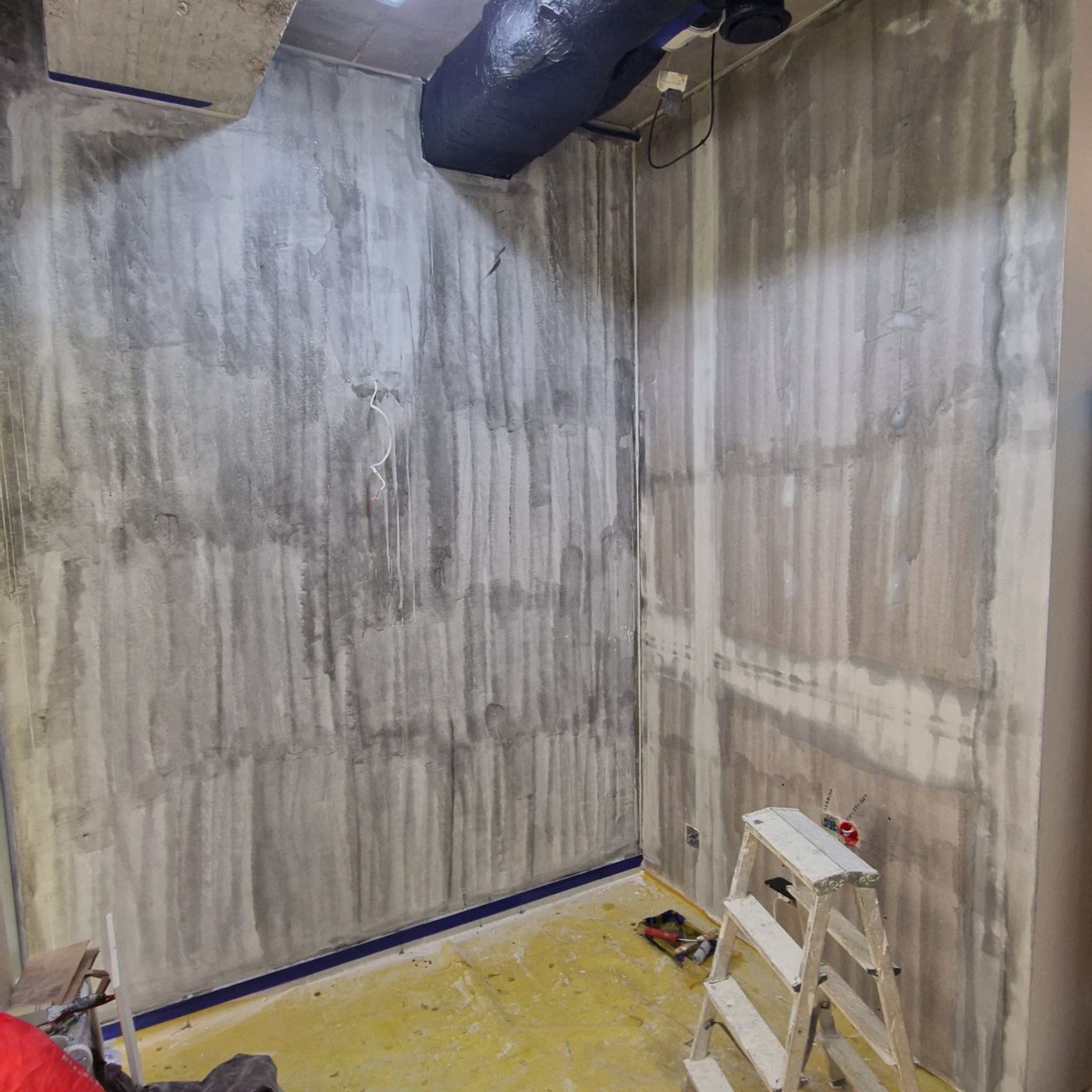 Pokój w trakcie nakładania gładzi, widoczne ślady po wałku, zabezpieczona taśmą podłoga, drabina przystawna w rogu pomieszczenia, widoczne wystające kable.
