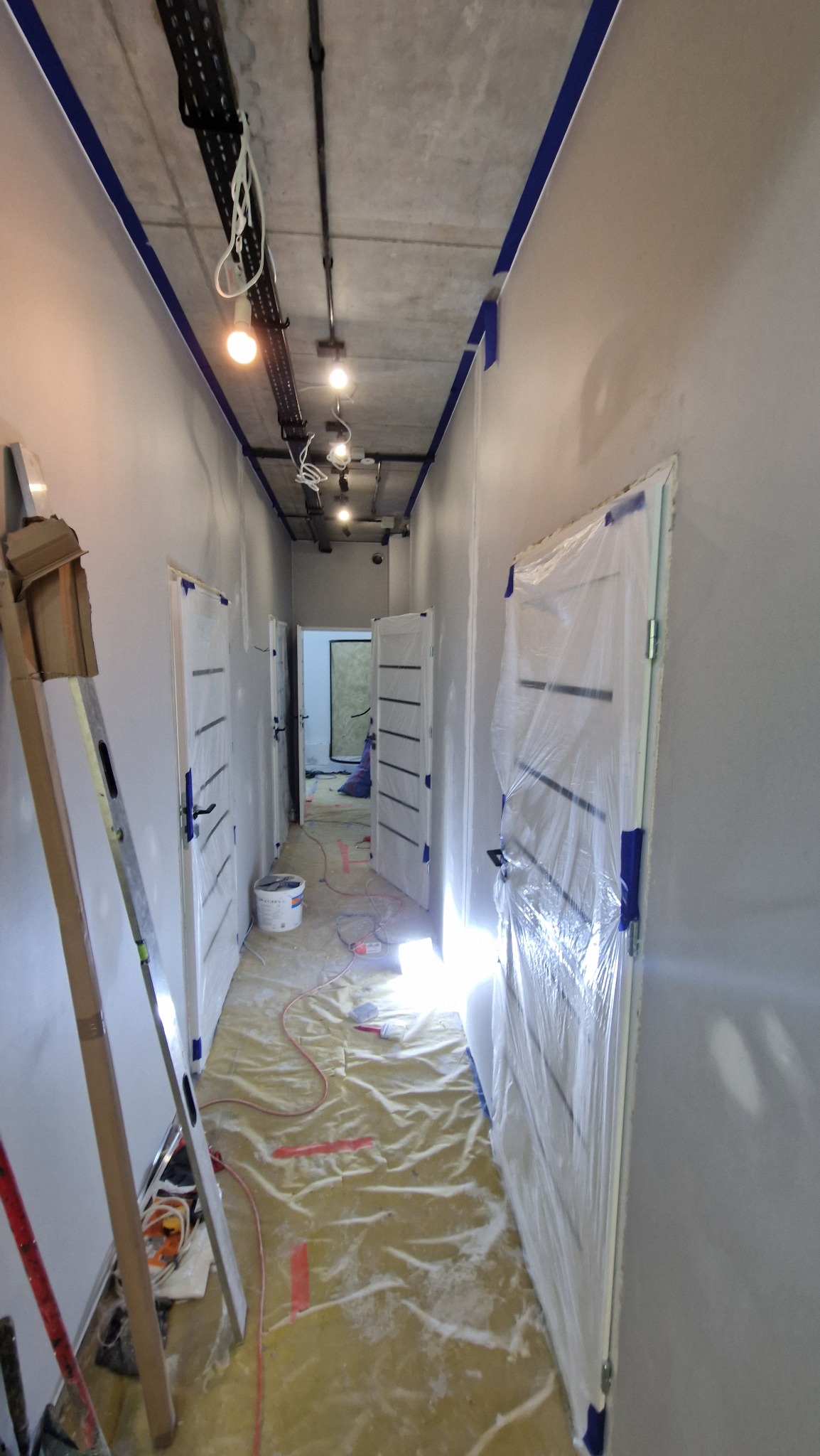 Korytarz w trakcie malowania, zabezpieczone folią drzwi, widoczna drabina, taśma malarska na suficie i ścianach, podłoga przykryta folią ochronną.