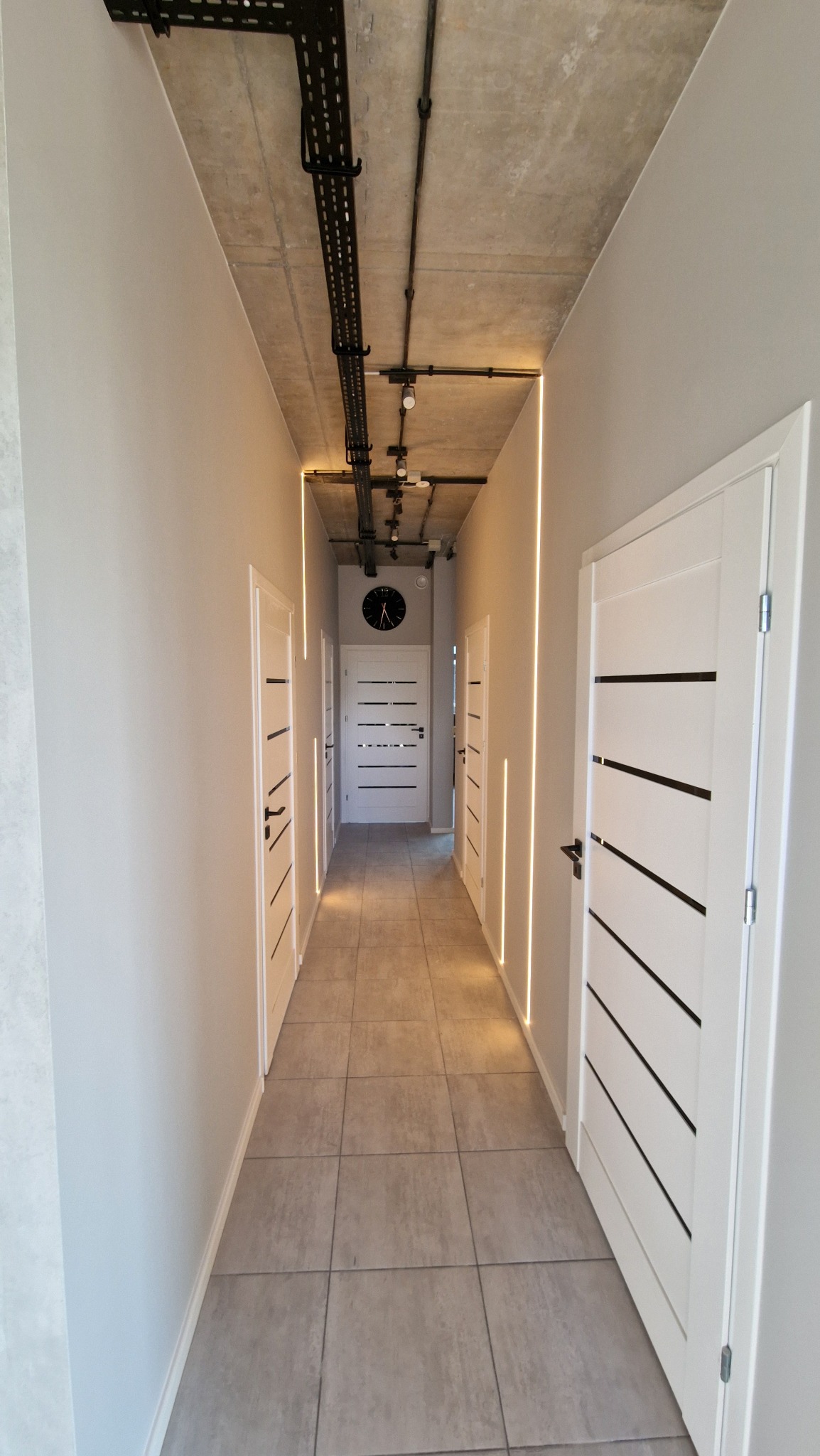 Nowoczesny korytarz z szarą podłogą z płytek, białymi drzwiami z czarnymi paskami, oświetleniem LED w pionowych wnękach i surowym betonowym sufitem z widoczną instalacją elektryczną.