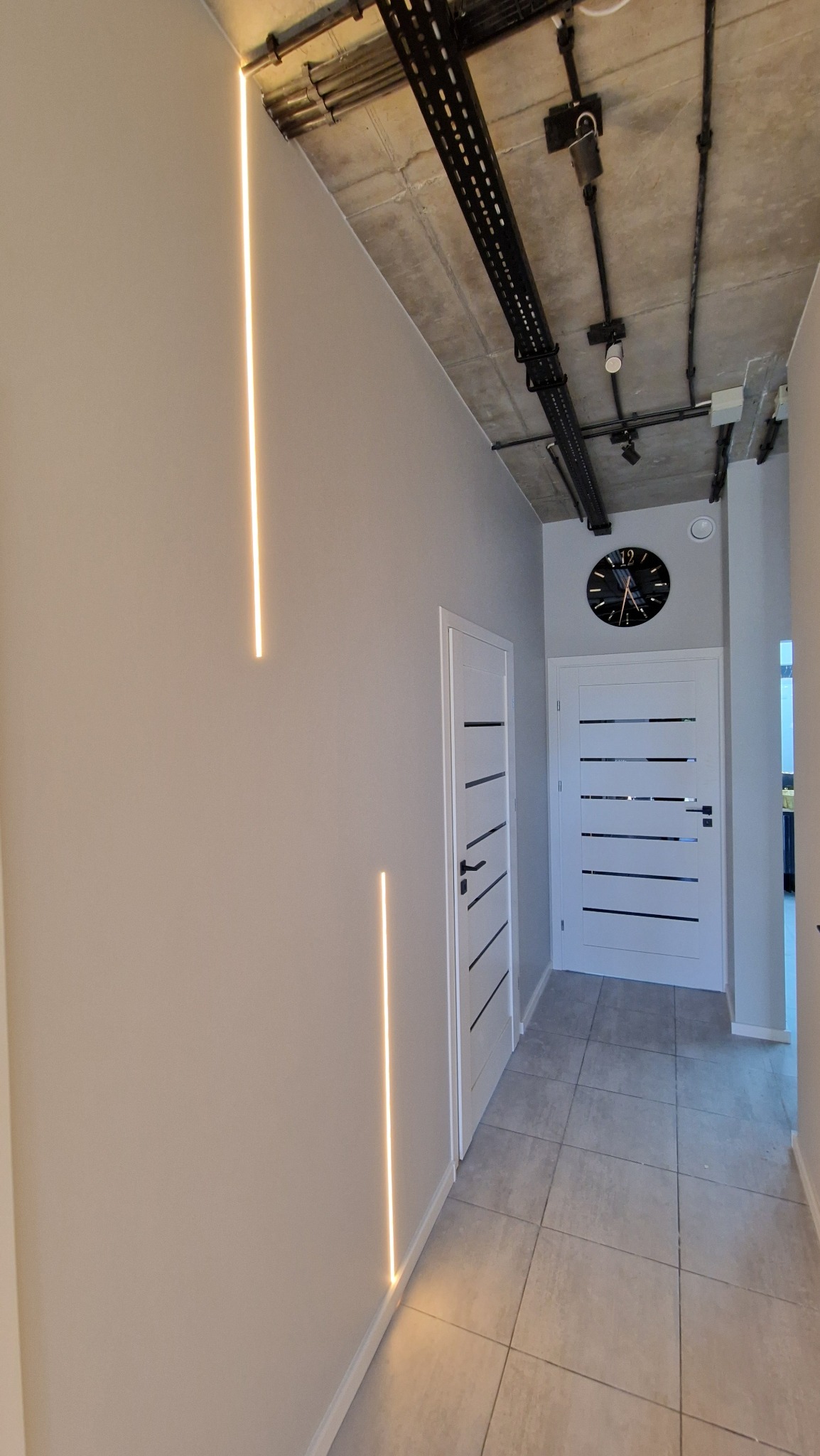 Nowoczesny korytarz z podświetlanymi pionowymi listwami LED na szarej ścianie, betonowy sufit z widocznymi instalacjami, dwie białe drzwi z czarnymi akcentami i duży zegar na ścianie.