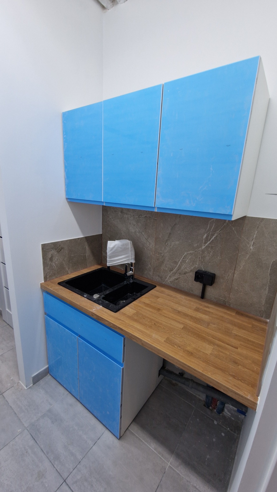 Niebieskie szafki kuchenne z blatem drewnopodobnym i czarnym zlewem, bateria zakryta białą szmatką, płytki imitujące marmur na ścianie, instalacja hydrauliczna widoczna pod blatem, folia ochronna...