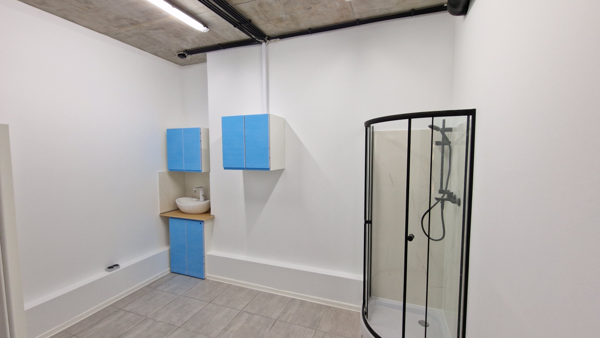 Nowoczesna łazienka z kabiną prysznicową z czarnymi ramami, umywalką nablatową i błękitnymi szafkami, surowy betonowy sufit z widocznymi instalacjami.