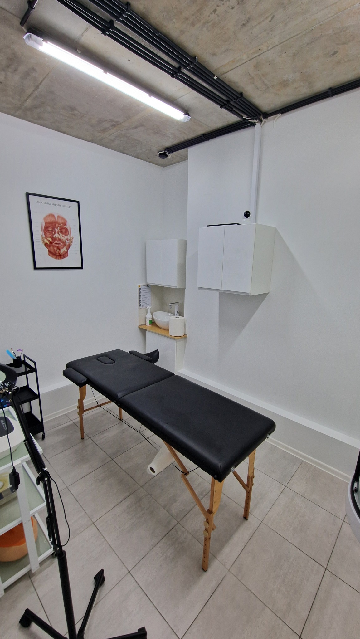 Klimatyczne wnętrze gabinetu kosmetycznego z czarnym leżakiem, umywalką i szafkami wiszącymi na białej ścianie, uzupełnione plakatem z anatomią mięśni twarzy.