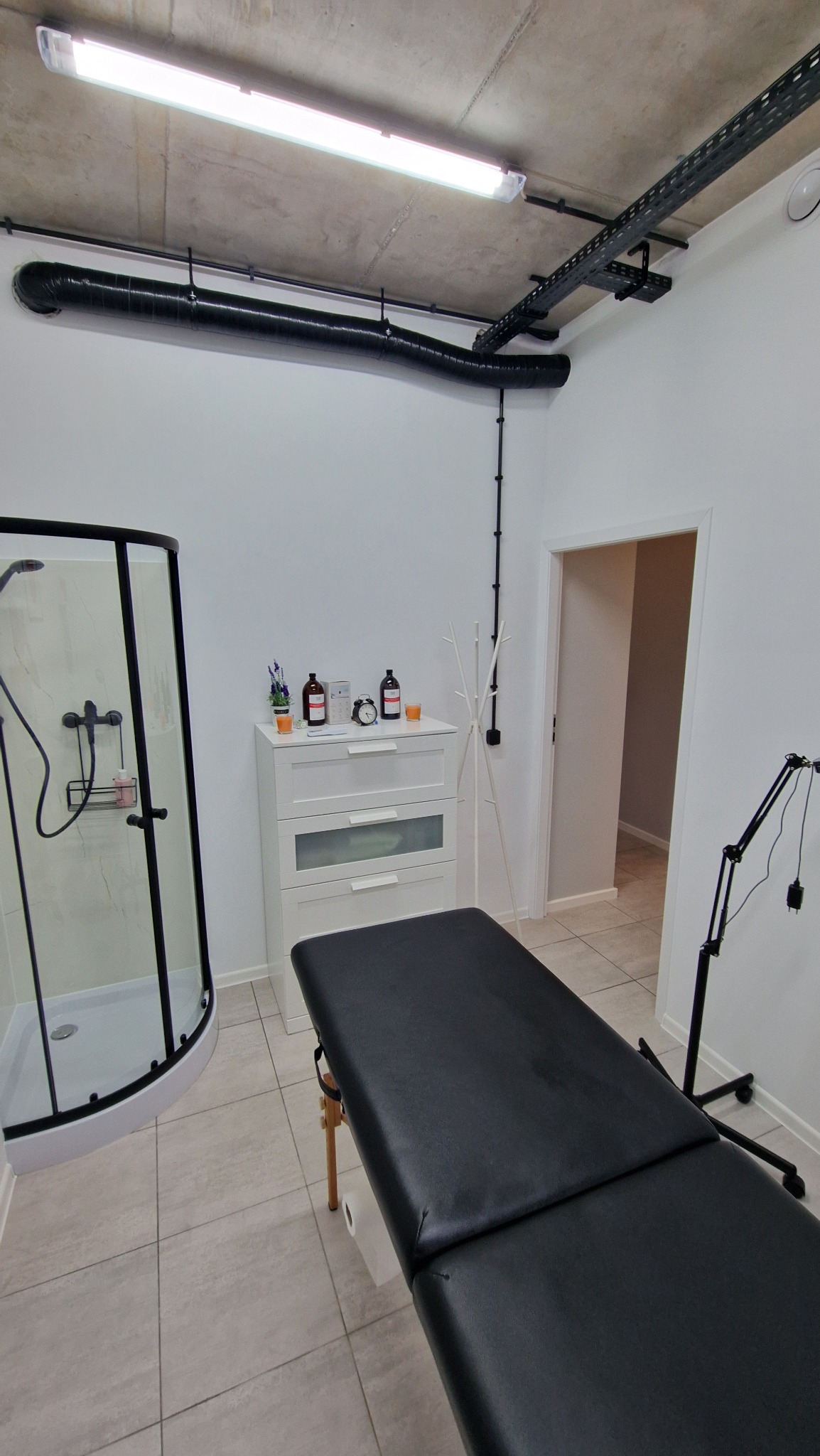 Nowoczesna łazienka z kabiną prysznicową, czarnym leżakiem do masażu, białą komodą i surowym betonowym sufitem z widocznymi instalacjami.