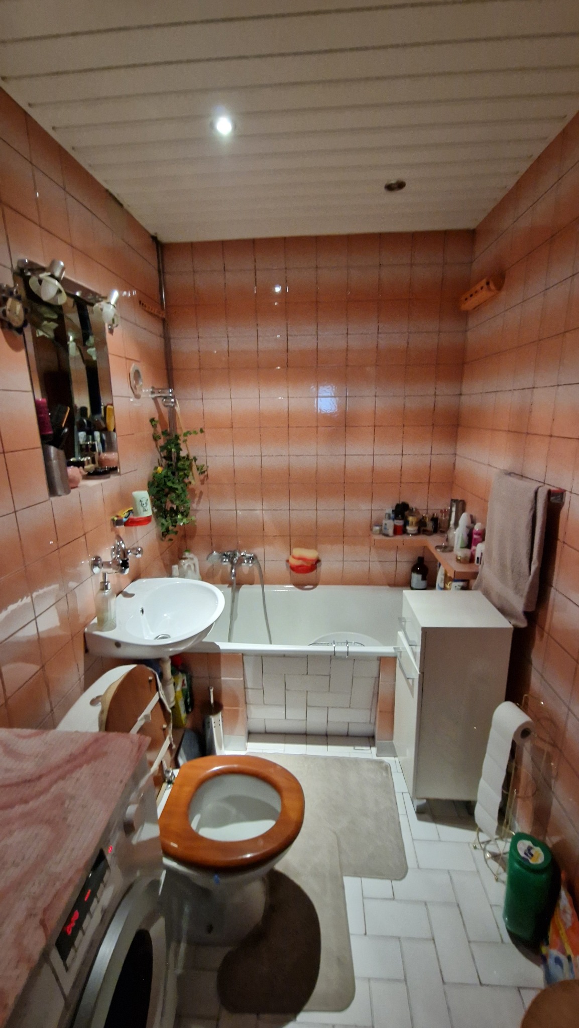 Łazienka w pomarańczowych płytkach z wanną, umywalką, toaletą z drewnianą deską i pralką z marmurowym blatem, widok na całe pomieszczenie.