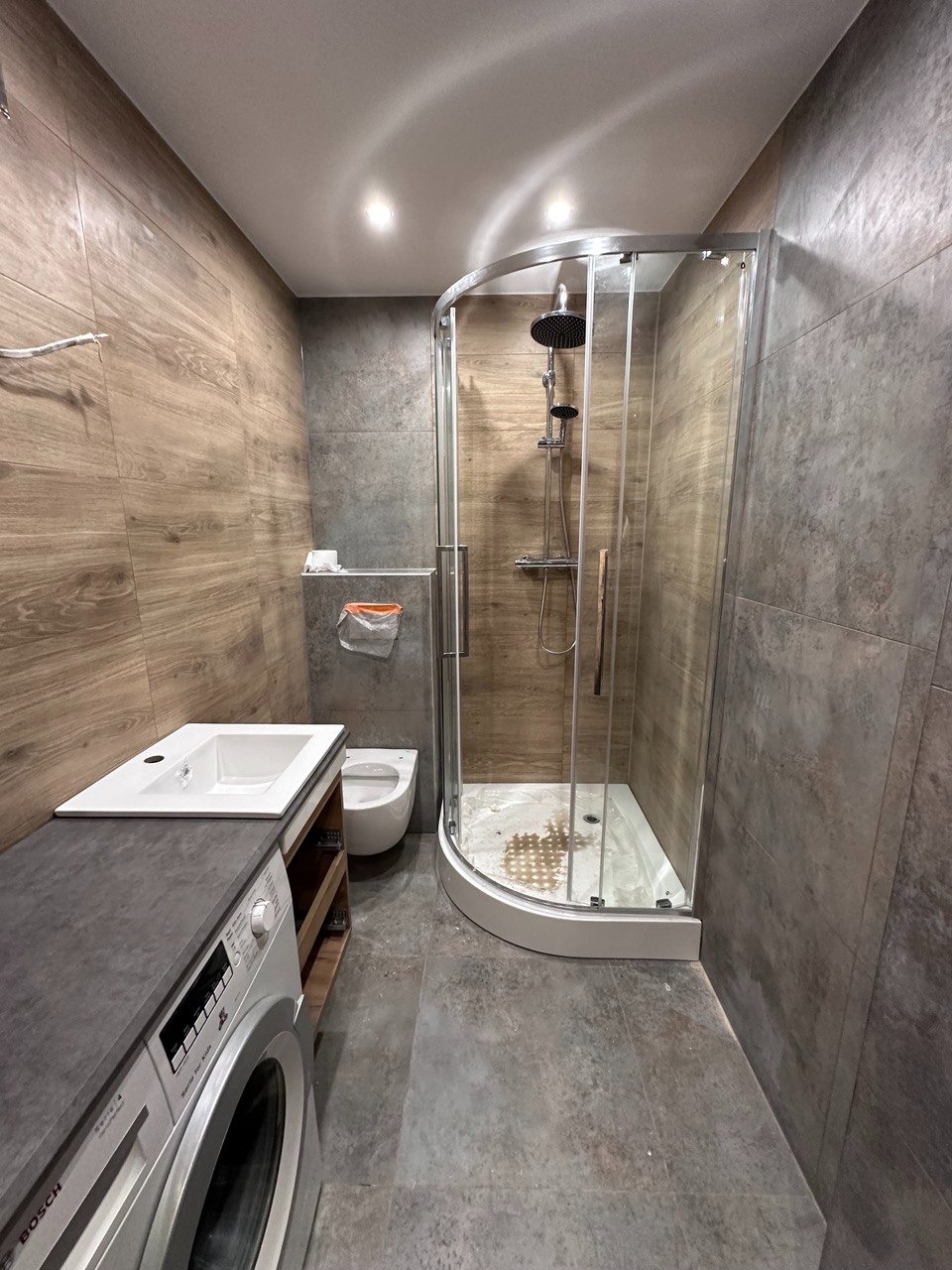 Nowoczesna łazienka z kabiną prysznicową typu walk-in, umywalką nablatową z szarą szafką i pralką Bosch Serie 4, wykończona płytkami imitującymi drewno i szarym betonem.