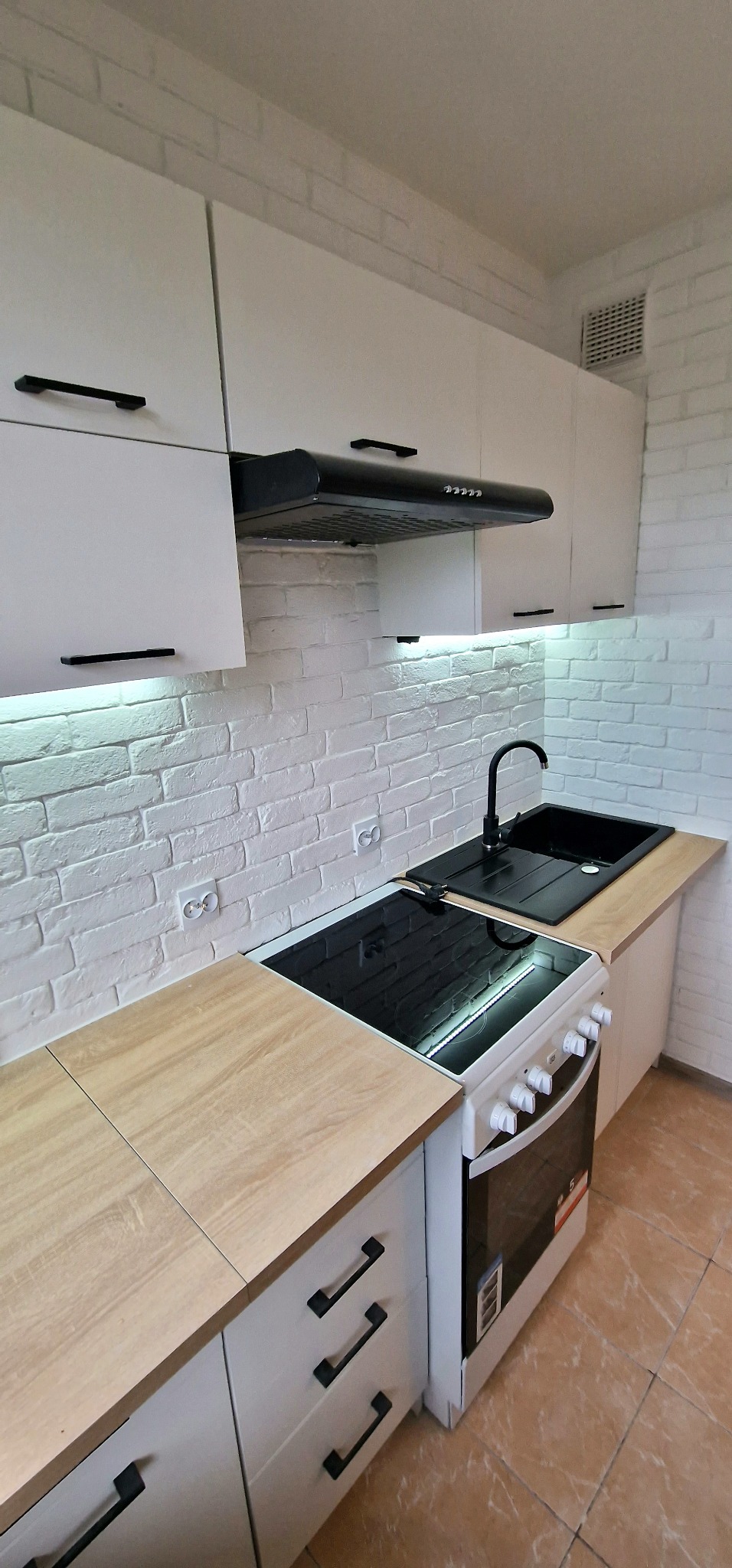 Nowoczesna kuchnia z białymi szafkami i czarnymi uchwytami, biała ceglana ściana, czarny zlew i bateria, płyta indukcyjna z piekarnikiem, oświetlenie LED pod szafkami.