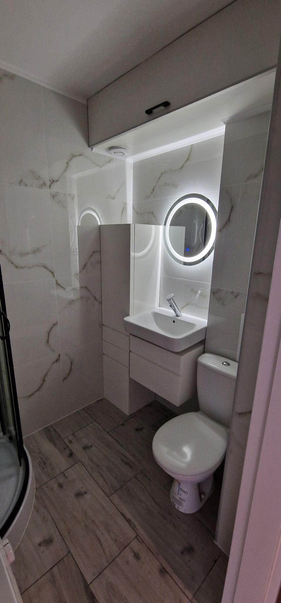 Nowoczesna łazienka z białymi płytkami imitującymi marmur, drewnopodobną podłogą, okrągłym lustrem z podświetleniem LED, wiszącą szafką z umywalką i toaletą marki Kolo.