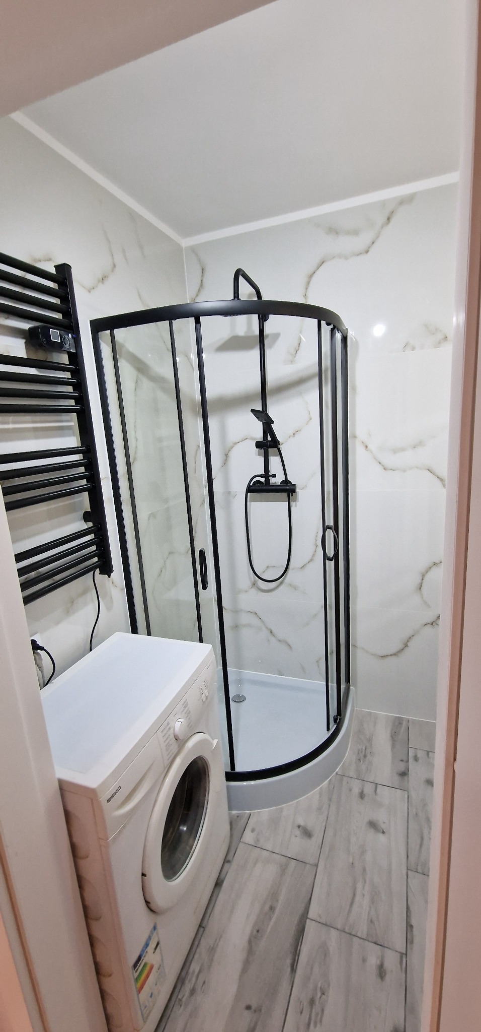 Nowoczesna łazienka po remoncie z czarną kabiną prysznicową, grzejnikiem drabinkowym i pralką. Na ścianach białe płytki z marmurkowym wzorem, na podłodze szare płytki imitujące drewno.