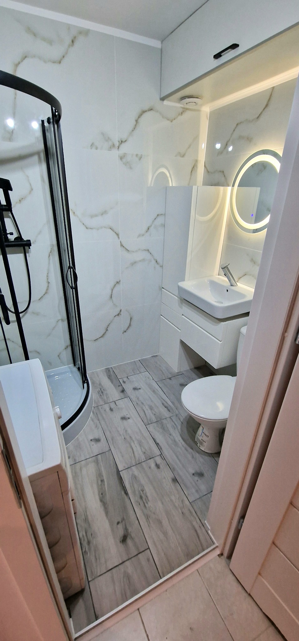 Nowoczesna łazienka z kabiną prysznicową z czarnymi ramami, umywalką na szafce i okrągłym lustrem z podświetleniem LED, wykończona płytkami imitującymi marmur i drewnopodobnymi płytkami podłogowymi.