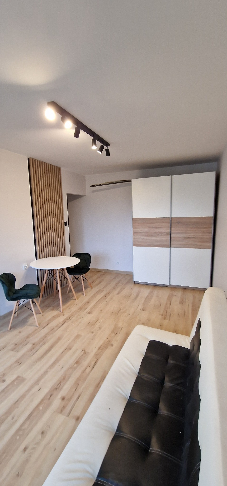 Jasne wnętrze pokoju z drewnianą podłogą, białym stolikiem i zielonymi krzesłami, biało-brązową szafą przesuwną oraz czarną listwą oświetleniową z reflektorami.