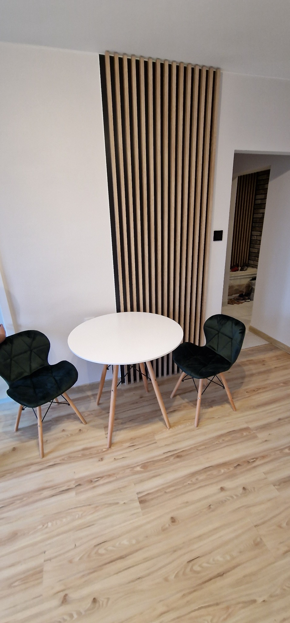 Nowoczesne wnętrze z drewnianym panelem lamelowym na ścianie, okrągłym białym stołem i dwoma ciemnozielonymi krzesłami w stylu skandynawskim na jasnej podłodze imitującej drewno.