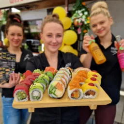 Trzy uśmiechnięte kelnerki prezentują drewnianą tacę z różnorodnym sushi i napoje w szklanych butelkach, w tle dekoracje z żółtych balonów i kwiatów.
