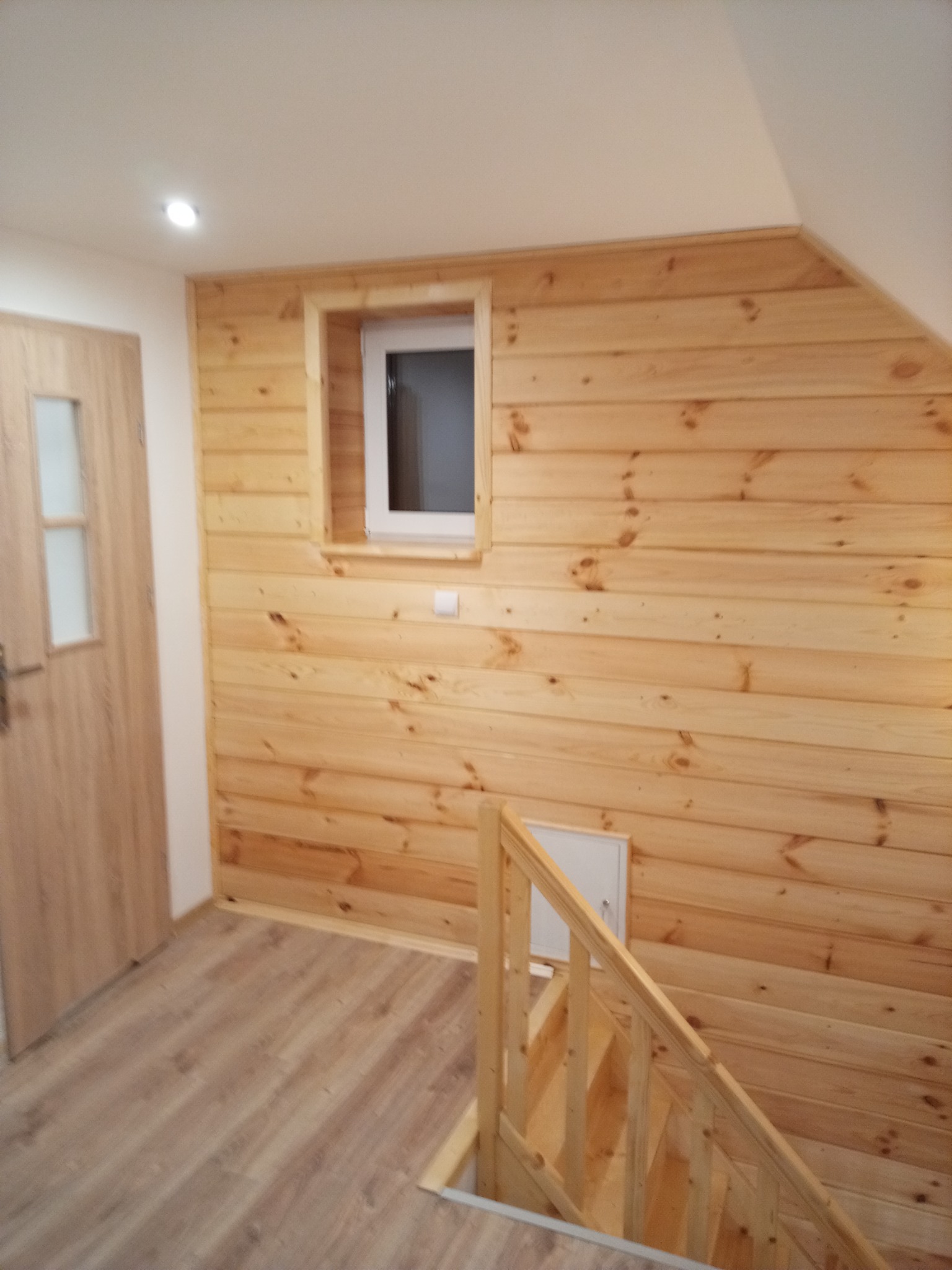 Drewniana boazeria ścienna z małym oknem i drzwiami, fragment schodów z drewnianą barierką, podłoga wyłożona panelami w odcieniach beżu.