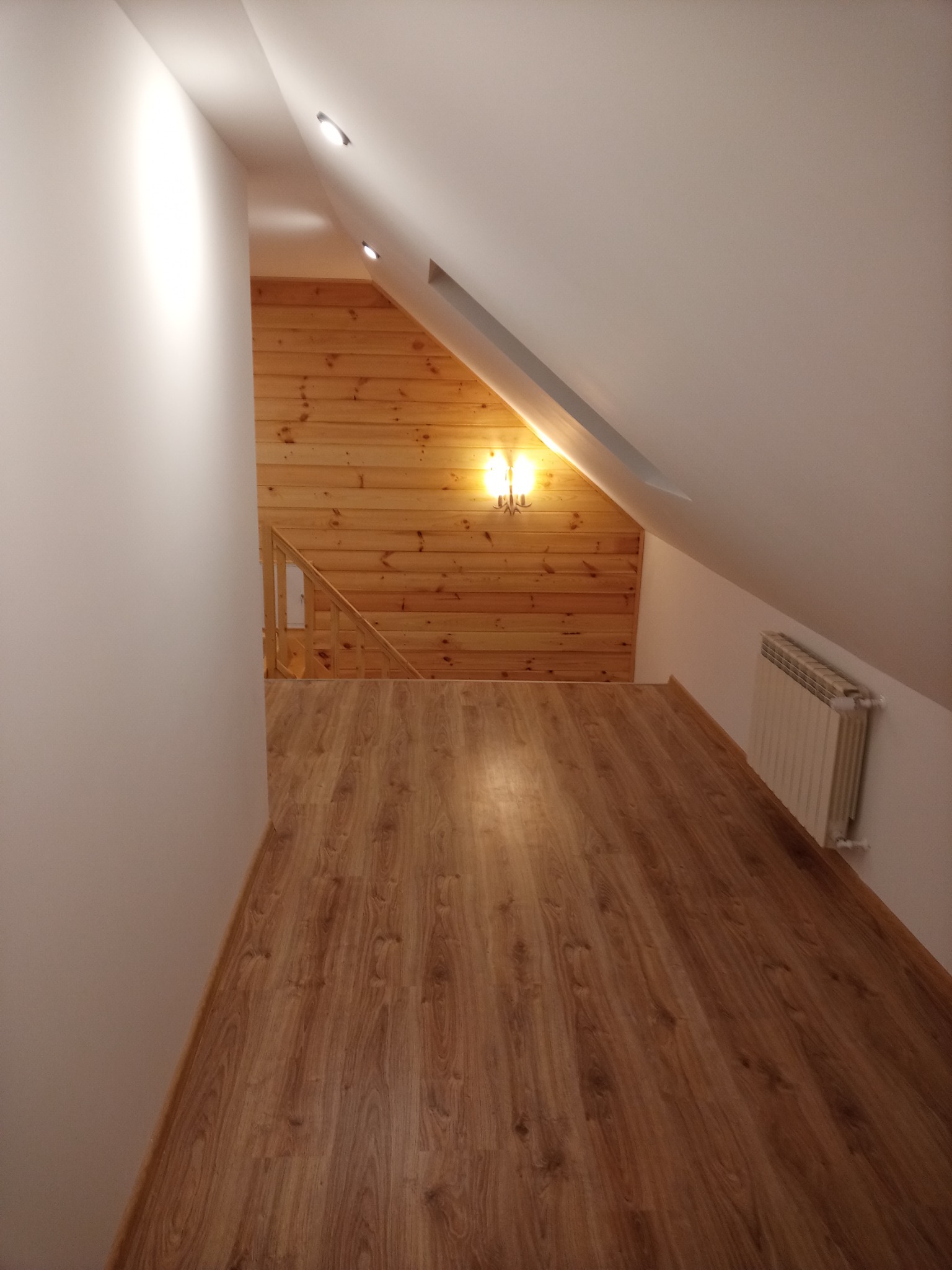 Wykończone poddasze z drewnianą boazerią, panele podłogowe w odcieniach brązu, grzejnik na białej ścianie i oświetlenie punktowe w skosach sufitu.